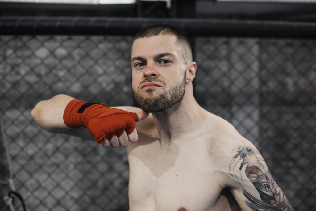 A muscular shirtless boxer with tattoos performing a chin flick gesture inside a cage.